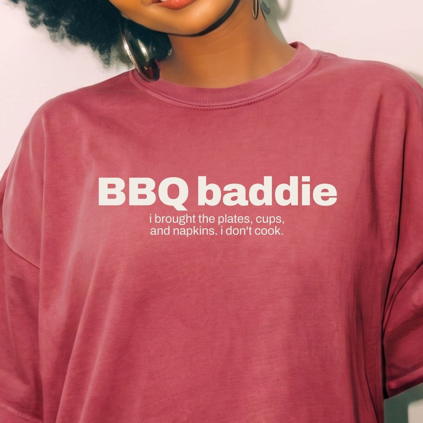 A woman with a black afro hairstyle wearing a relaxed-fit, faded burgundy t-shirt with white text that reads 'BBQ baddie' and a smaller humorous definition below, set against a plain light background.