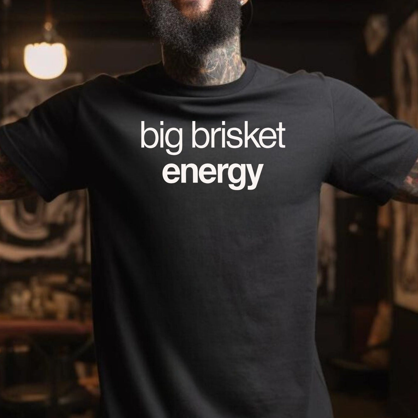 A man with a beard and tattoos wearing a black t-shirt with the text 'big brisket energy' in white, standing in a dimly lit bar or cafe.