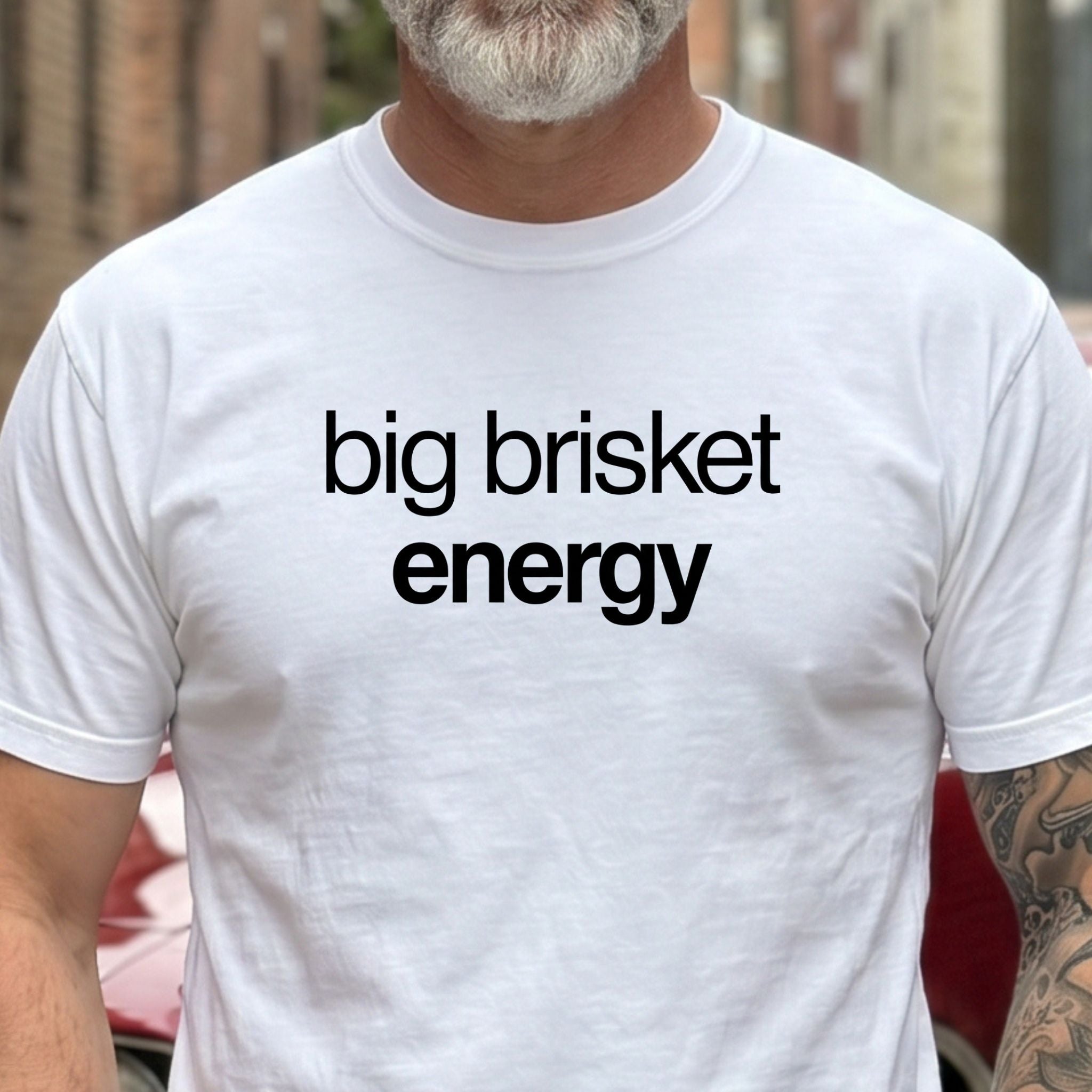 A man wearing a white crewneck t-shirt with the phrase 'big brisket energy' printed in black lowercase letters across the chest, set against a blurred outdoor background.