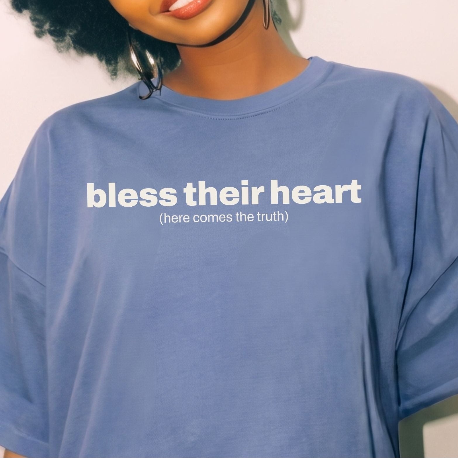 A woman with curly black hair wearing a blue oversized t-shirt with the white printed text 'bless their heart (here comes the truth)'.