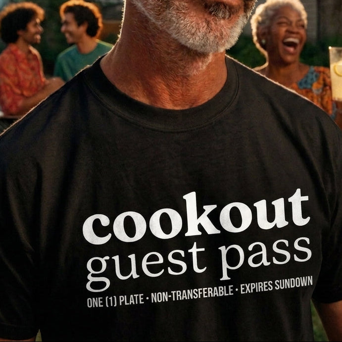 A man wearing a black graphic t-shirt with a white 'cookout guest pass' print, standing in a backyard party setting with string lights and other people blurred in the background.