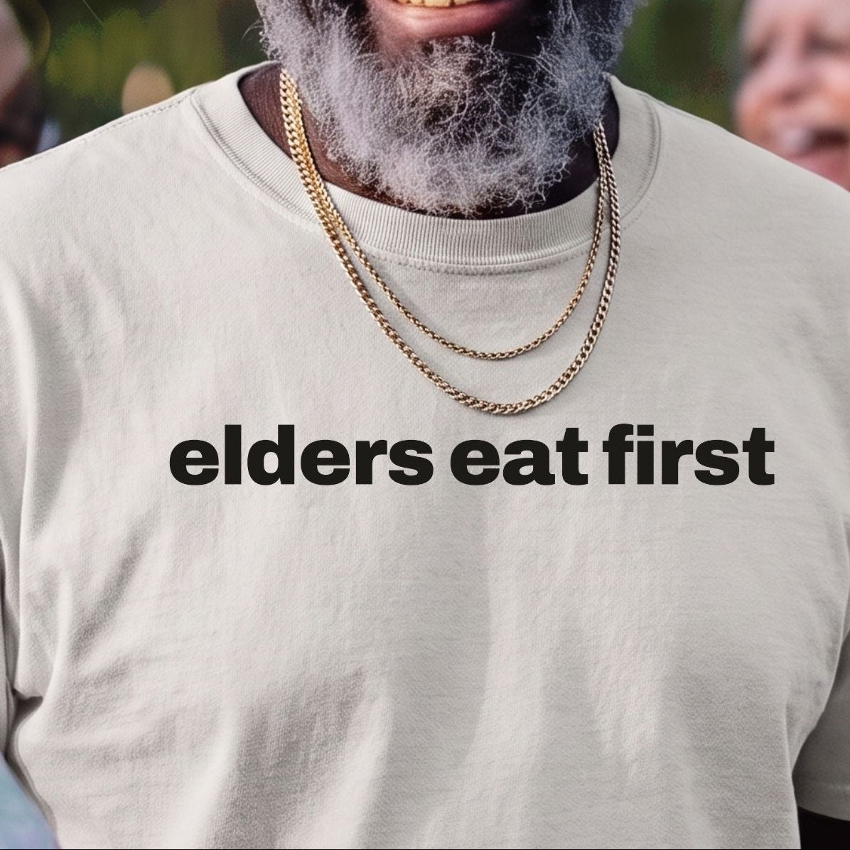 A smiling older man outdoors wearing a straw hat, dark sunglasses, layered gold necklaces, and a light beige T-shirt with the phrase "elders eat first" printed in black across the chest. Other people are partially visible in the blurred background.