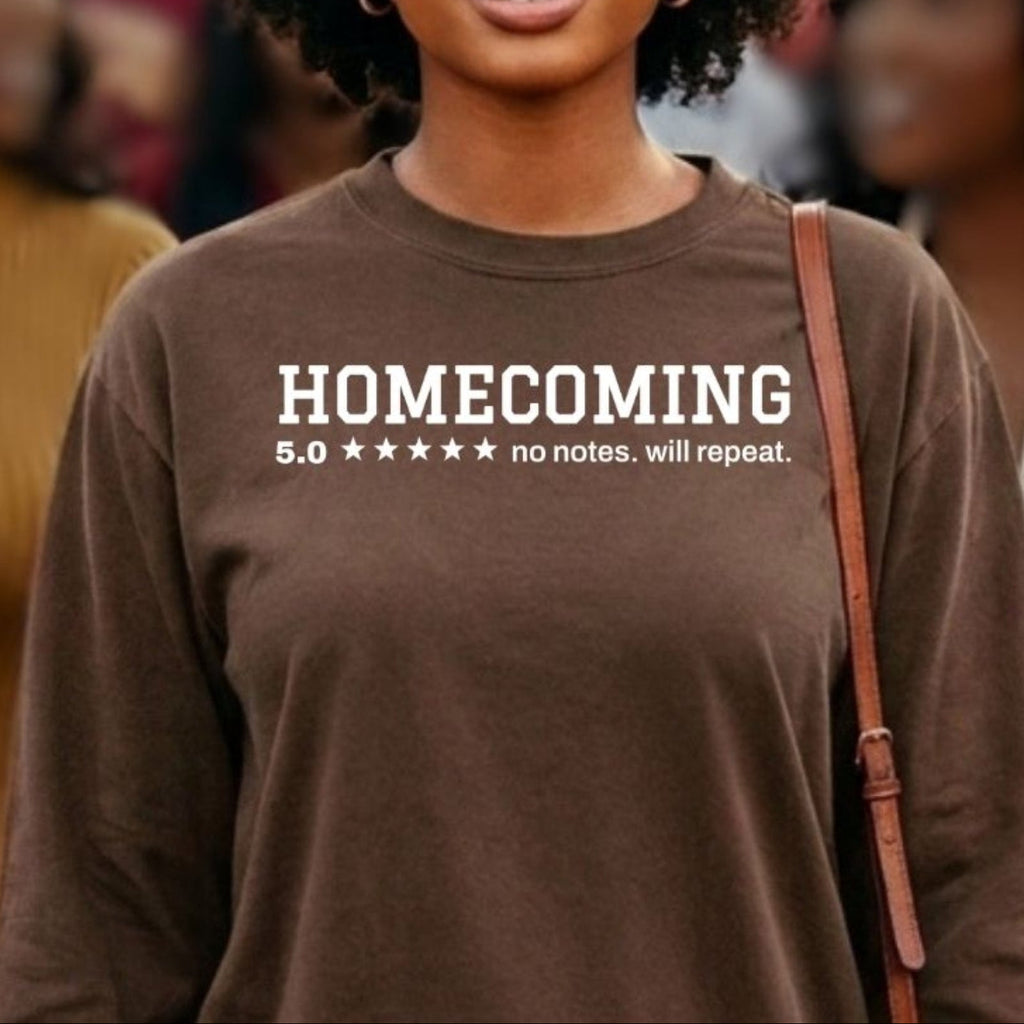 A woman wearing a brown long-sleeve t-shirt with white printed text that reads 'HOMECOMING 5.0 ***** no notes, will repeat.