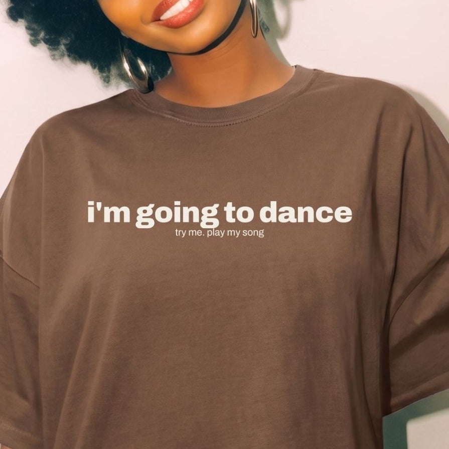 A woman with a large black afro is wearing an oversized brown t-shirt with the white printed text 'i'm going to dance' and smaller text 'try me, play my song'.