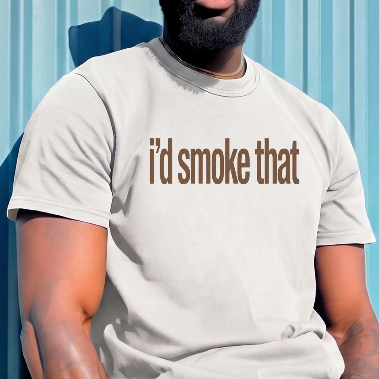 A man wearing a light beige bucket hat, sunglasses, and a matching light beige t-shirt with the text 'i'd smoke that' printed in brown across the chest, sitting against a blue corrugated metal wall.