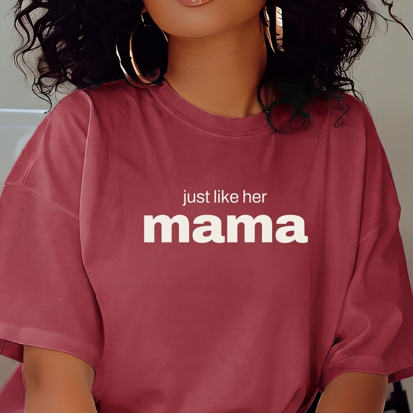 A woman with voluminous curly black hair wearing a dusty rose pink oversized t-shirt with the white printed text 'just like her mama'. She is wearing large gold hoop earrings and posing indoors.