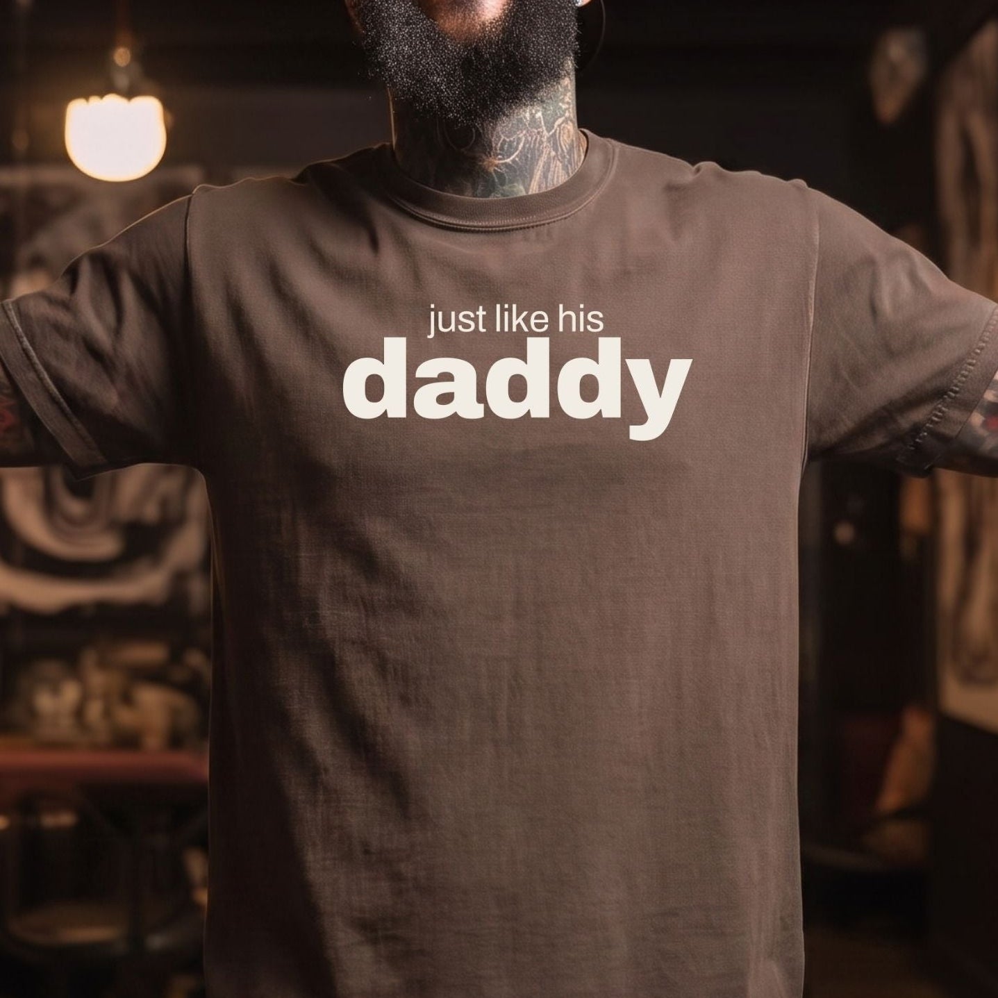 A smiling man with a beard and tattoos wearing a dark brown t-shirt with the text 'just like his daddy' printed in white across the chest, standing in a dimly lit indoor environment.