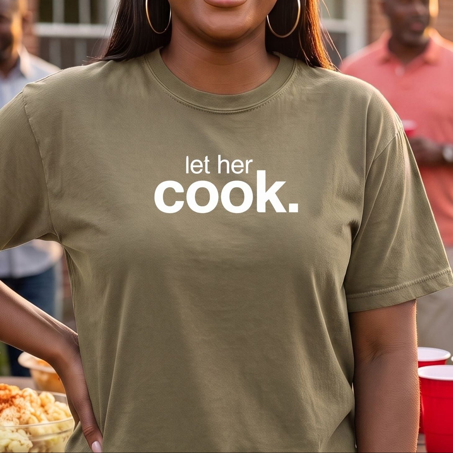 A woman smiling outdoors at a backyard BBQ wearing an olive green t-shirt with the white printed text 'let her cook.'