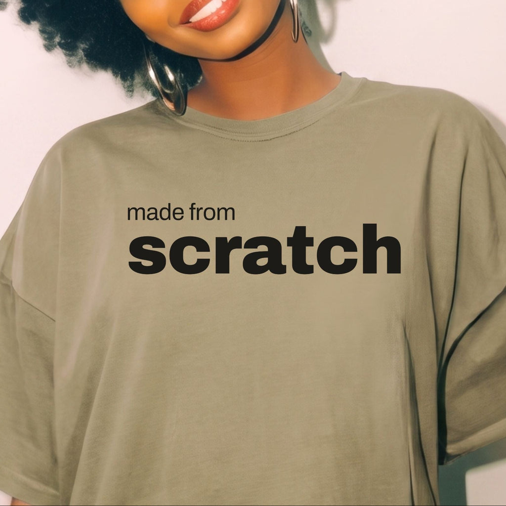 A woman with a large black afro is wearing an oversized olive green t-shirt with the text 'made from scratch' printed in black across the chest. She is wearing large silver hoop earrings and smiling against a plain off-white background.
