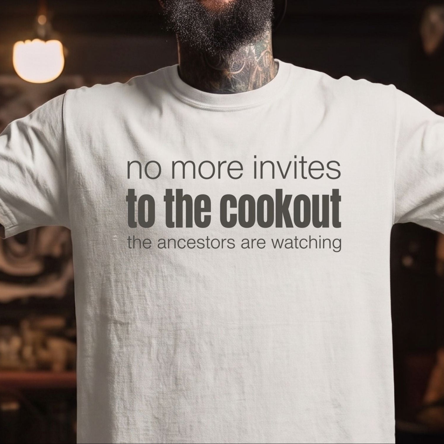 A man with a beard and tattoos wearing a white graphic t-shirt and a black baseball cap, smiling with arms outstretched in a dimly lit indoor setting. The t-shirt features the text 'no more invites to the cookout the ancestors are watching' in dark gray.