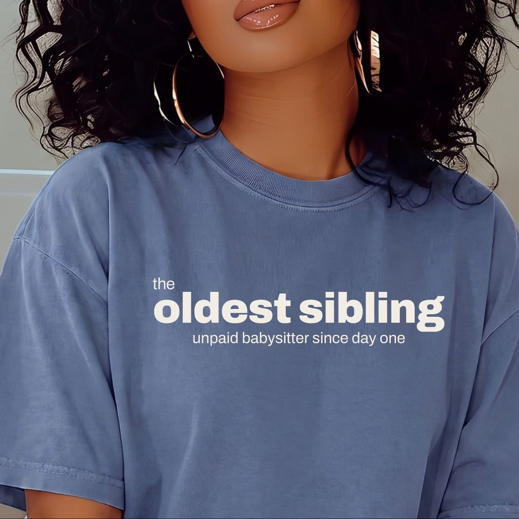 A woman with voluminous curly black hair wearing a light blue oversized t-shirt with the text 'the oldest sibling unpaid babysitter since day one' in white. She is also wearing large gold hoop earrings.