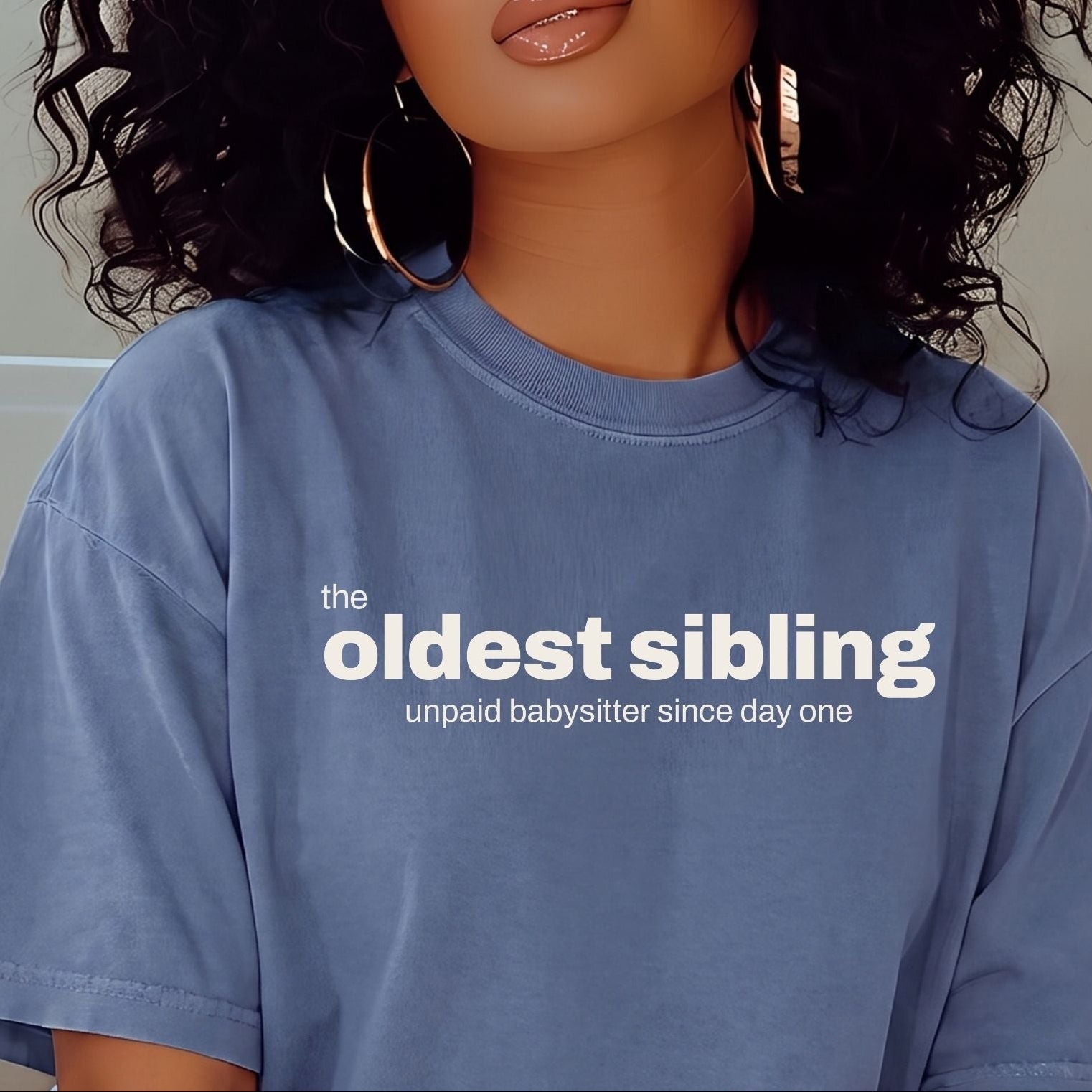 A woman with voluminous curly black hair wearing a light blue oversized t-shirt with the text 'the oldest sibling unpaid babysitter since day one' in white. She is also wearing large gold hoop earrings.