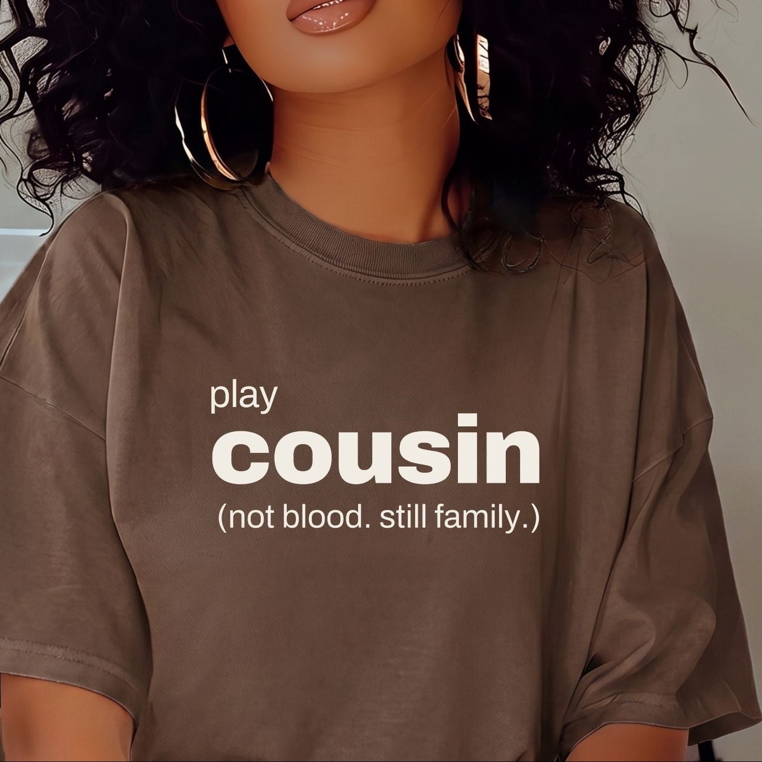 A woman with voluminous curly black hair wearing a brown graphic t-shirt with the text 'play cousin (not blood. still family.