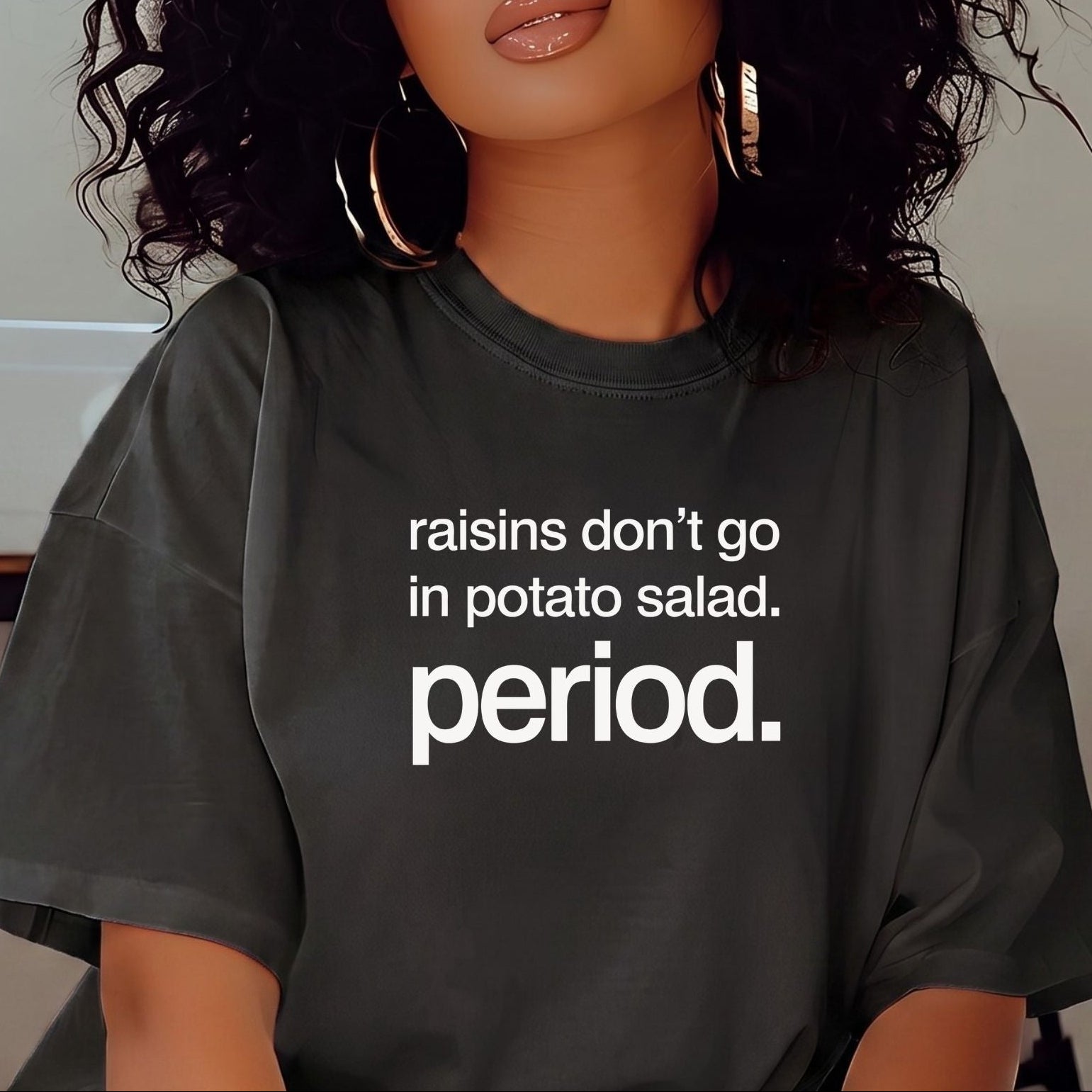 A woman with voluminous dark curly hair is wearing a dark gray oversized t-shirt with the white printed text 'raisins don't go in potato salad. period.
