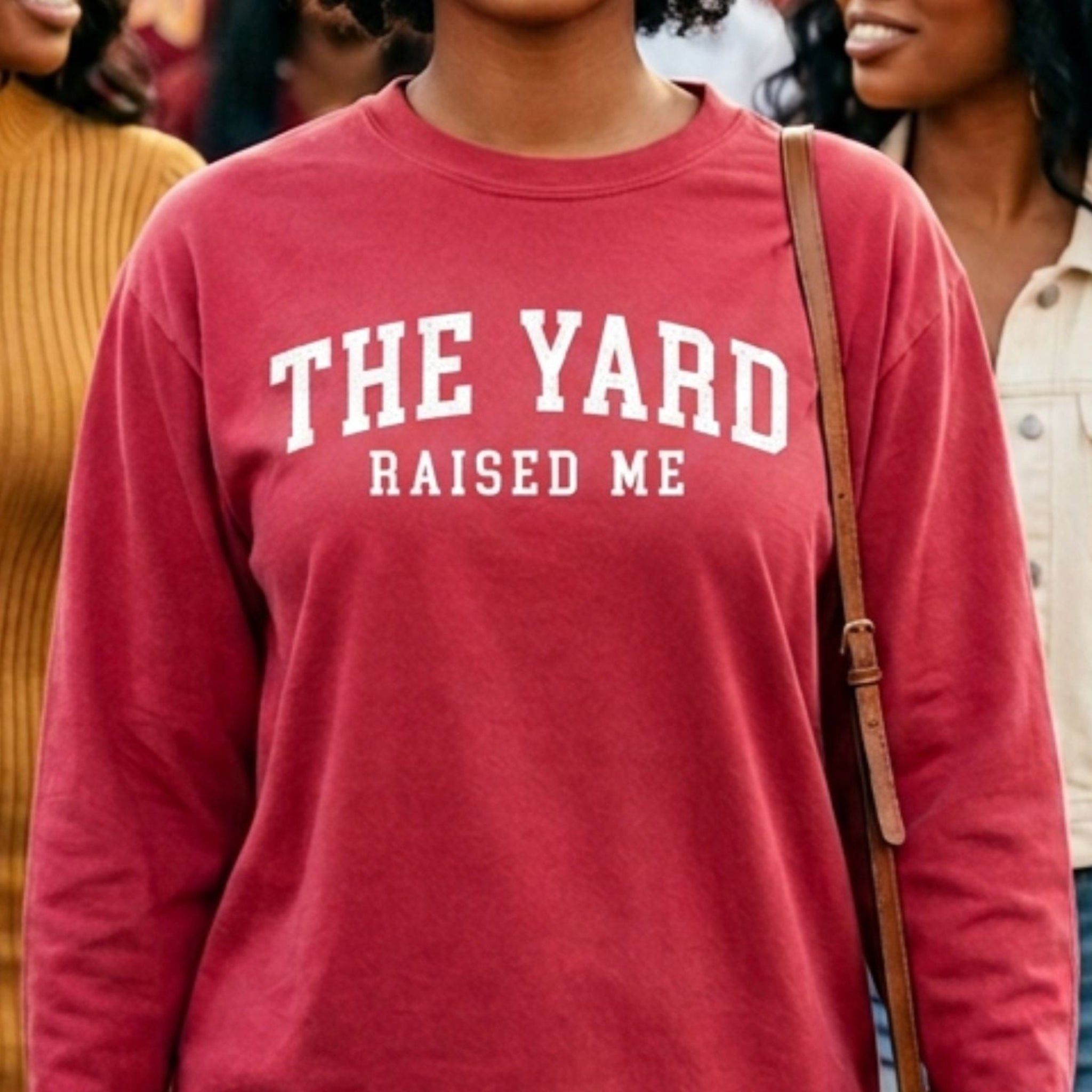 A person wearing a faded red long-sleeve crewneck sweatshirt with white text reading 'THE YARD RAISED ME'. The photo is cropped from the neck down, showing the person wearing light blue jeans and carrying a brown shoulder bag, with other people partially visible in the background.