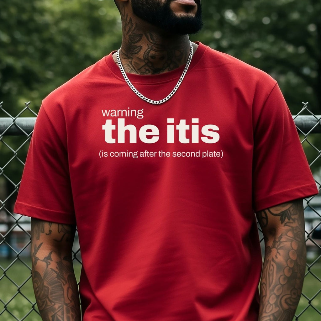 A man wearing a red graphic t-shirt with white text that reads 'warning the itis (is coming after the second plate)' stands in front of a chain-link fence with green trees in the background.