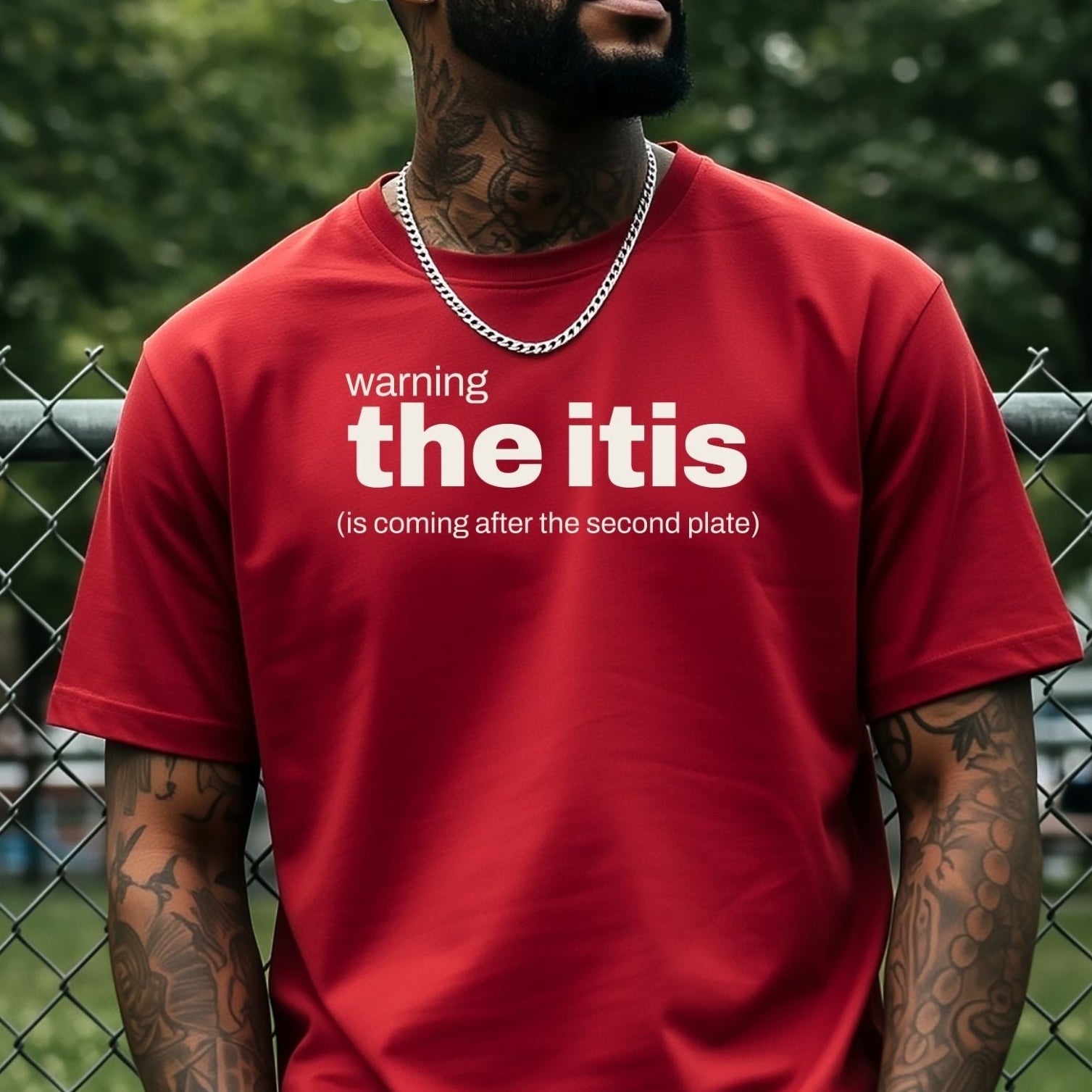 A man wearing a red graphic t-shirt with white text that reads 'warning the itis (is coming after the second plate)' stands in front of a chain-link fence with green trees in the background.