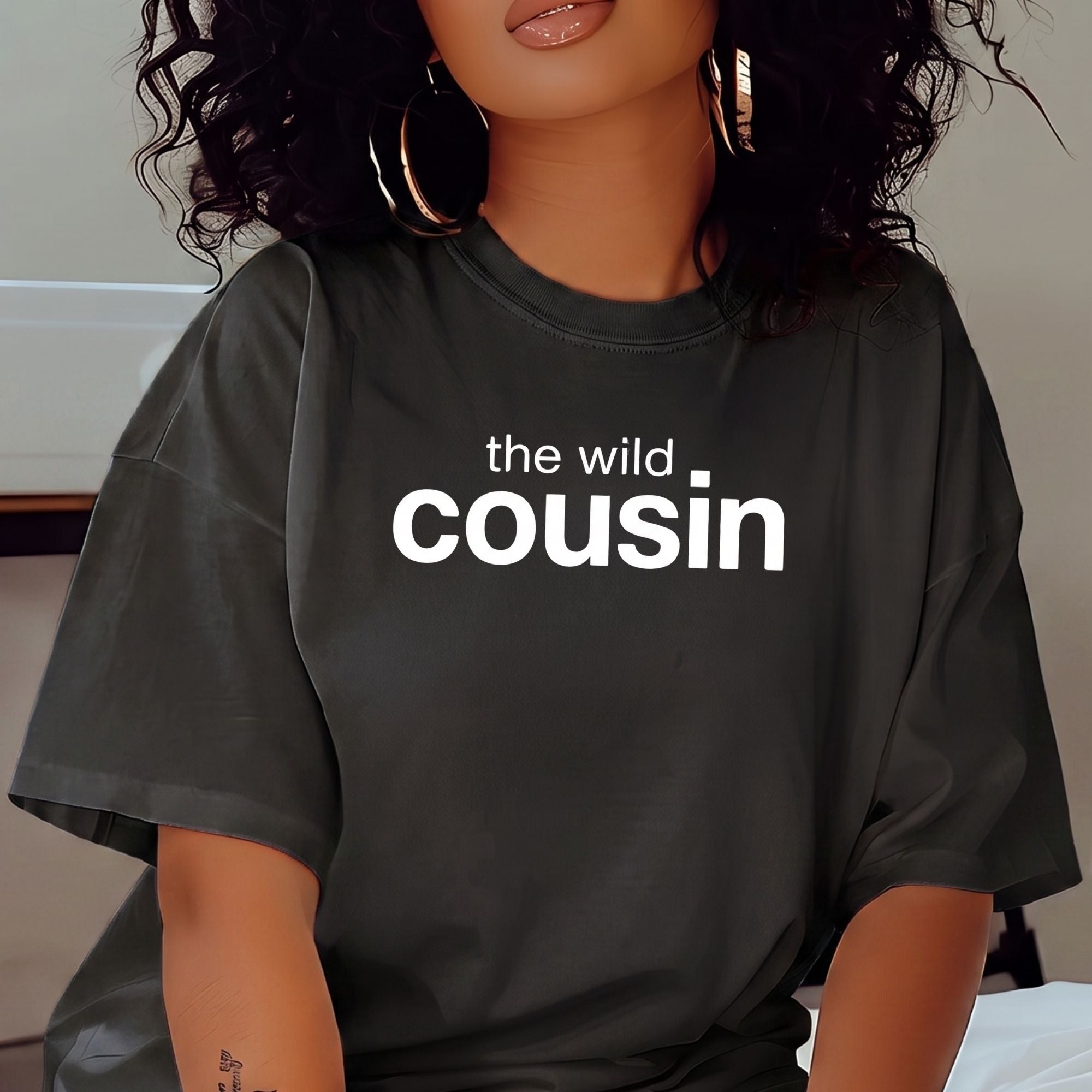 A woman wearing an oversized dark charcoal gray t-shirt with the phrase 'the wild cousin' printed in white across the chest, styled with large gold hoop earrings.