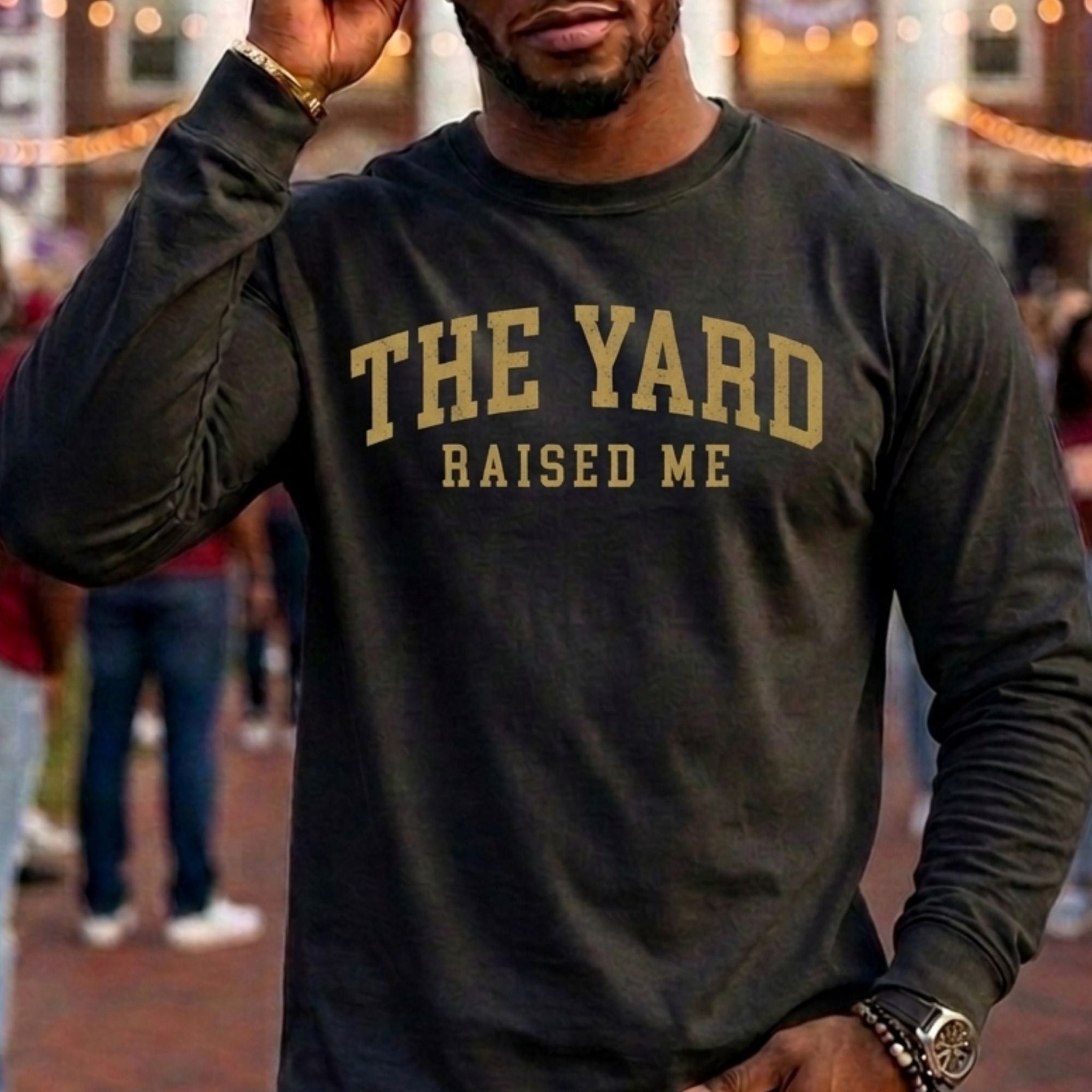 A man wearing a dark charcoal long-sleeve t-shirt with 'THE YARD RAISED ME' printed in gold across the chest, set against a blurred outdoor background with warm lights and people.