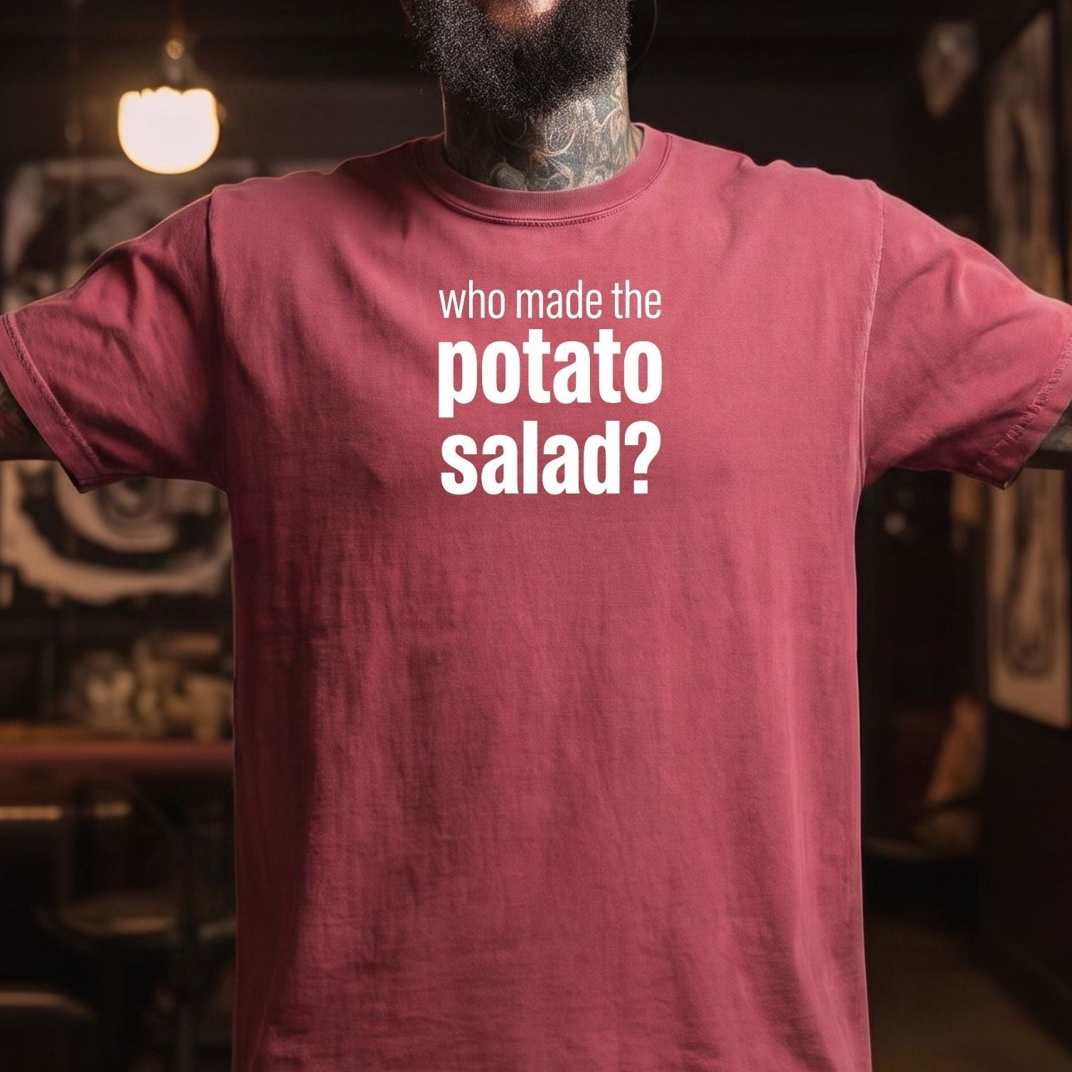 A smiling man with a beard and tattoos wearing a maroon t-shirt with the white text 'who made the potato salad?' on the chest, standing in a dimly lit indoor environment.